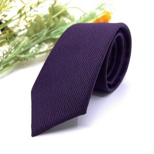 May include: A deep purple necktie featuring a subtle, diagonal ribbed texture throughout the fabric. The tie is rolled at one end, showcasing the consistent weave pattern and the rich, dark violet color of the material from the pointed tip to the roll.