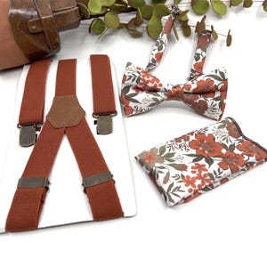 May include: A set of rust-colored suspenders with brown leather accents, a floral bow tie, and a matching pocket square. The bow tie and pocket square feature a white background with orange and green floral designs.