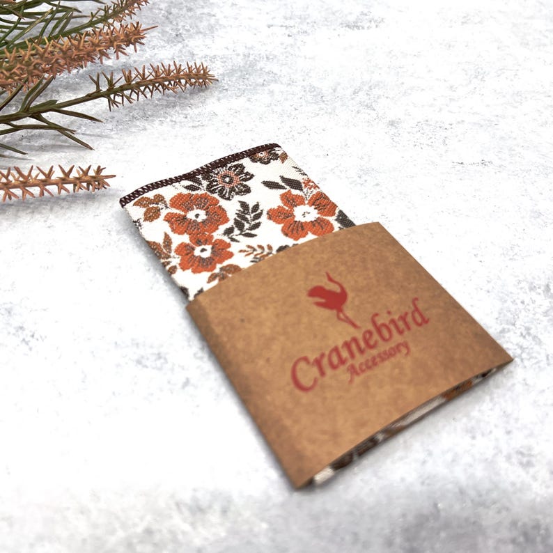 May include: A folded pocket square featuring a white background with a repeating floral pattern in shades of orange and dark brown. The fabric is partially encased in a brown paper sleeve printed with the red text Cranebird Accessory.