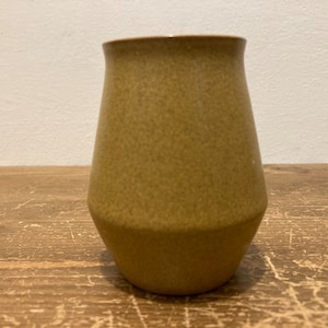 May include: A small, light brown ceramic vase with a textured surface. The vase has a rounded bottom and a slightly flared opening.