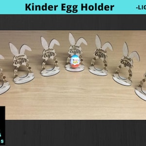 May include: Six wooden bunny-shaped holders for Kinder Eggs. Each holder has the text "Happy Easter" on it. One holder has a Kinder Egg inside.