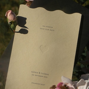 Könnte beinhalten: Eine beigefarbene Save-the-Date-Karte mit einem Herz-Ausschnitt und dem Text "Wir heiraten Save Our Date Sophie & Thomas 09. September 2023 Einladung folgt". Die Karte ist von rosa Rosen umgeben.