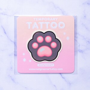 Può includere: Un tatuaggio temporaneo rosa e grigio a forma di impronta di zampa di gatto. Il tatuaggio è su uno sfondo bianco con il testo "TEMPORARY TATTOO" e "natgreenart" sulla carta.