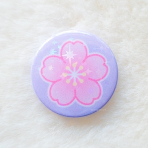May include: A purple button featuring a pink cherry blossom design. The flower has a white centre and is surrounded by small white stars.