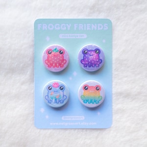 Könnte beinhalten: Ein Set mit vier Mini-Abzeichen mit Frosch-Motiven. Die Abzeichen sind rund und zeigen verschiedene farbige Frösche mit unterschiedlichen Designs. Die Abzeichen sind auf einem blauen Hintergrund in einem Raster angeordnet, mit dem Text "Froggy Friends mini badge set" und "www.notgreenart.etsy.com".