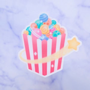 May include: A pink and white striped popcorn bucket filled with colorful planets and a yellow shooting star. The planets have rings and are various shades of blue, green, pink, and orange.