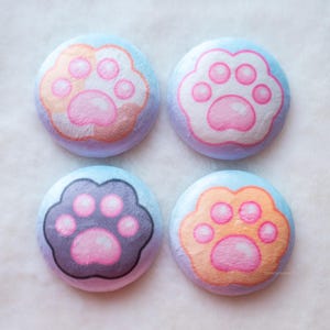 Pode incluir: Quatro botões redondos com pegadas de gato. Cada botão tem um fundo azul claro e uma pegada em tons de rosa, laranja e cinza. Os botões estão dispostos em um quadrado sobre uma superfície branca e fofa.