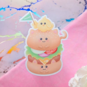 May include: A cute cartoon sticker of a vegan burger with a cat face, topped with a chick and a flag that says "VEGAN".