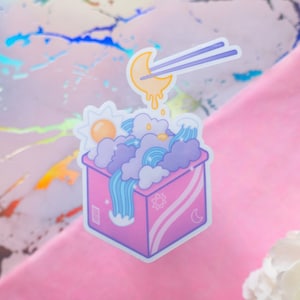 May include: A pink box with a blue, white, and yellow sky inside. The box has a sun and moon design on the side. A yellow crescent moon with a dripping yellow liquid is being lifted by chopsticks.