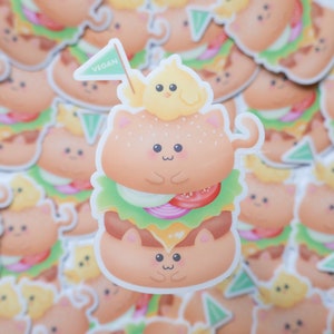 May include: A cute cartoon cat dressed as a vegan burger with a small yellow chick on top. The cat is wearing a green flag that says "VEGAN".