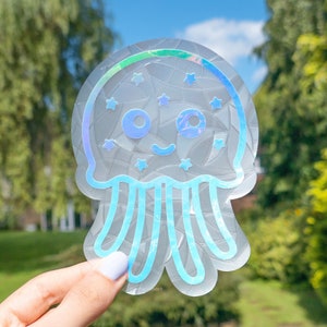 May include: A holographic sticker of a blue and white jellyfish with a smiling face and stars on its body. The sticker is iridescent and has a cracked glass effect.