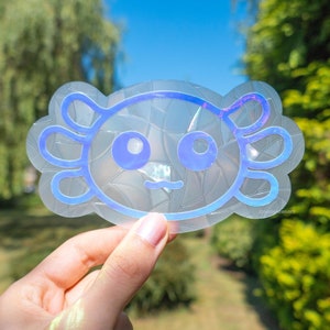 May include: A clear iridescent sticker with a blue outline of an axolotl. The axolotl has a simple face with two eyes and a small mouth.