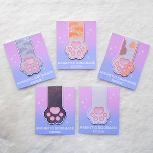 May include: Five different cat paw-shaped magnetic bookmarks with glitter accents. Each bookmark features a different cat paw print design with a pink, purple, and blue gradient background. The text "MAGNETIC BOOKMARK" is printed on each bookmark.