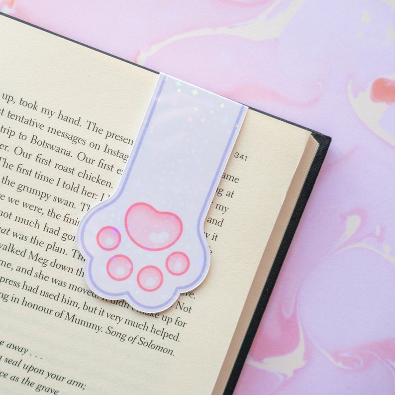 May include: A pink and white paw print bookmark with a glittery purple background and stars. The bookmark is made of a clear, durable material.