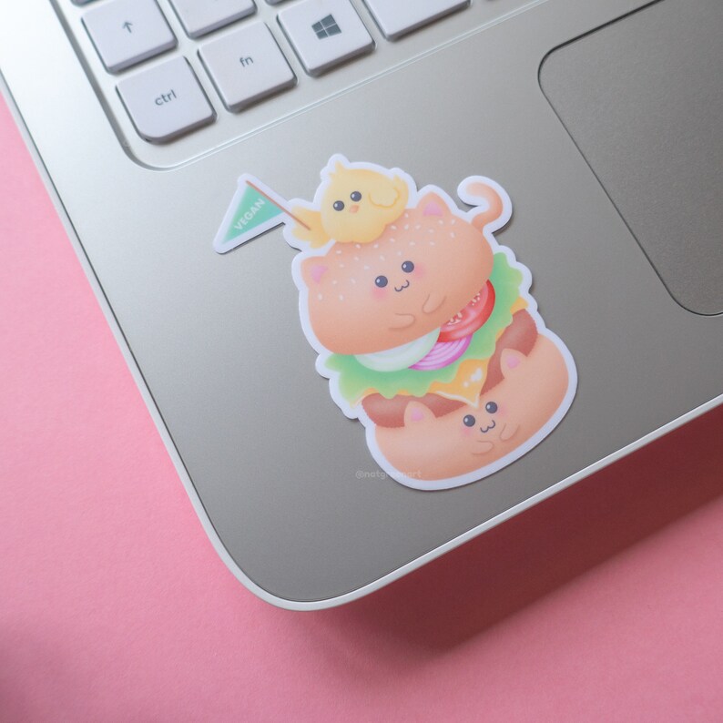May include: A sticker of a vegan burger made of cute cartoon cats. The burger is topped with lettuce, tomato, onion, and cheese. A small yellow chick is perched on top of the burger, holding a green flag that says "vegan".