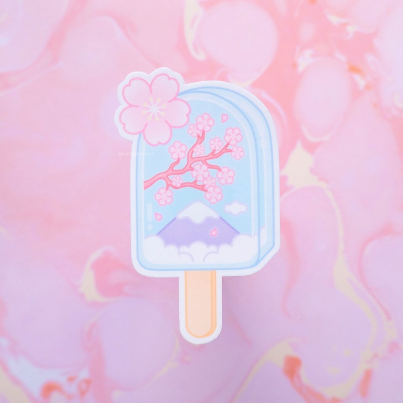 May include: A pastel blue popsicle sticker with a white stick. The popsicle features a pink cherry blossom branch, a purple mountain, and white clouds.