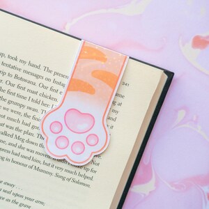 May include: A pink and orange cat paw bookmark with glitter accents. The bookmark is shaped like a paw print with four pink toes and a pink pad.