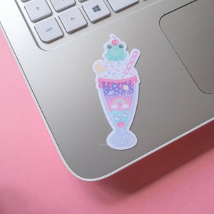May include: A colorful sticker featuring a whimsical milkshake design. The milkshake has a light blue base with a rainbow, planets, and a cute frog on top. The sticker is decorated with sprinkles, a cherry, and a straw.