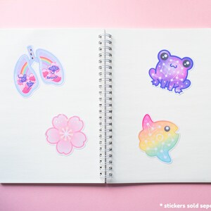 May include: A notebook with four colorful stickers on the pages. The stickers feature a pair of lungs, a flower, a frog, and a fish. Each sticker has a rainbow gradient and a constellation design.