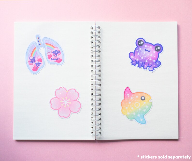 May include: A notebook with four colorful stickers on the pages. The stickers feature a pair of lungs, a flower, a frog, and a fish. Each sticker has a rainbow gradient and a constellation design.