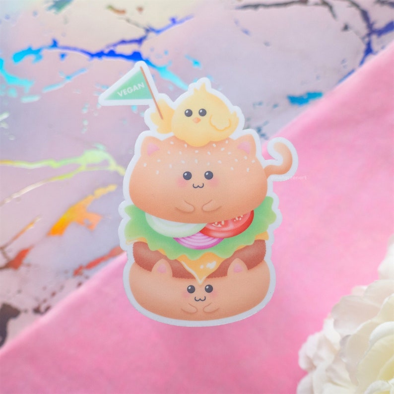 May include: A cute cartoon sticker of a vegan burger with a cat face, topped with a chick and a flag that says "VEGAN".