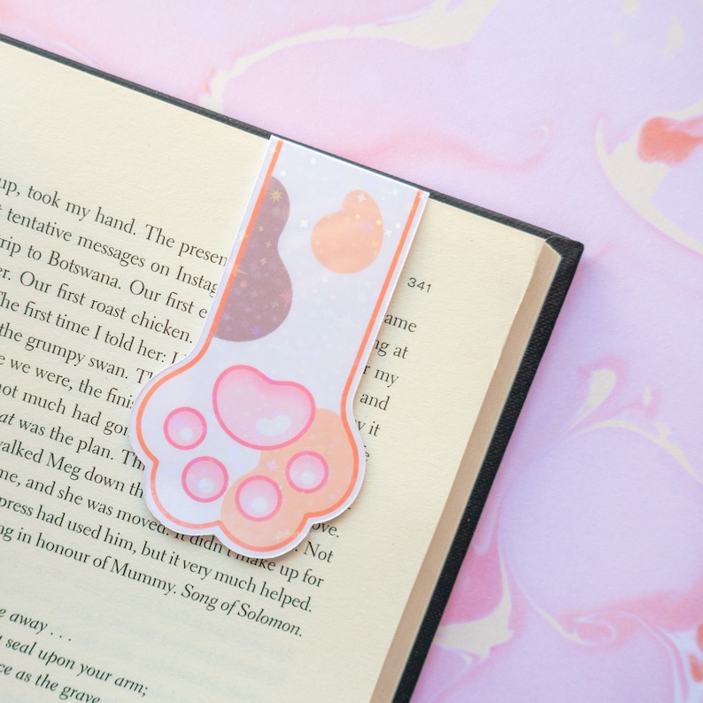 May include: A pink and orange paw print bookmark with glitter accents. The bookmark is shaped like a cat's paw with four toes and a pink pad. The background is a white book page with text.