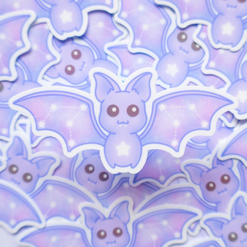 May include: A close-up of a stack of purple and white bat stickers with a constellation design. The bats have a cute cartoon style with big eyes and a star on their chest.