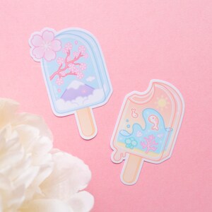 May include: Two colorful popsicle stickers with a summer theme. One sticker features a blue popsicle with a mountain, clouds, and pink cherry blossoms. The other sticker features a peach popsicle with a blue whale, coral, and bubbles.