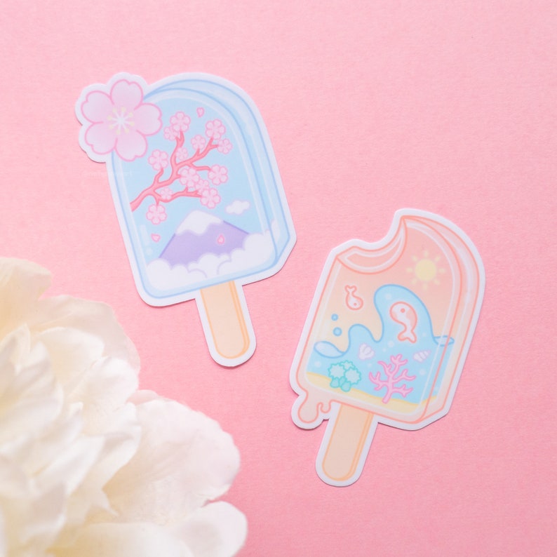 May include: Two colorful popsicle stickers with a summer theme. One sticker features a blue popsicle with a mountain, clouds, and pink cherry blossoms. The other sticker features a peach popsicle with a blue whale, coral, and bubbles.