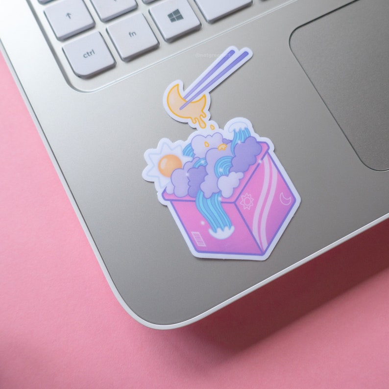 May include: A pink box with a white and blue design filled with clouds, a sun, a moon, and a wave. A pair of chopsticks are holding a yellow crescent moon with a white drip.