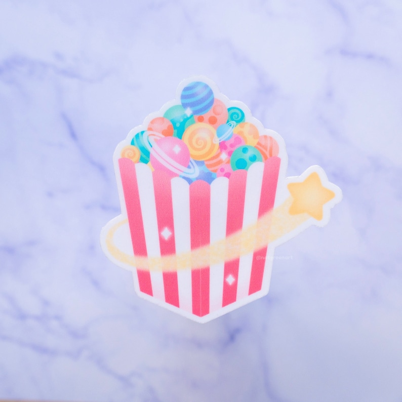 May include: A pink and white striped popcorn bucket filled with colorful planets and a yellow shooting star. The planets have rings and are various shades of blue, green, pink, and orange.