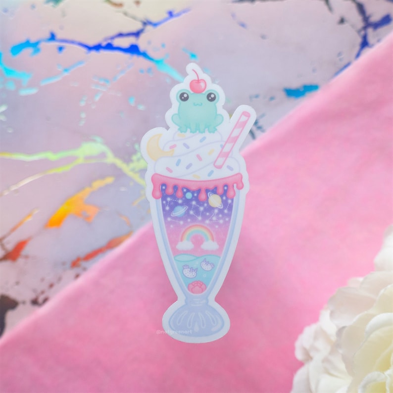 May include: A whimsical sticker featuring a milkshake with a frog on top. The milkshake has a pink and purple color scheme, with a rainbow, planets, and a cherry on top. The sticker is a cute and colorful design.