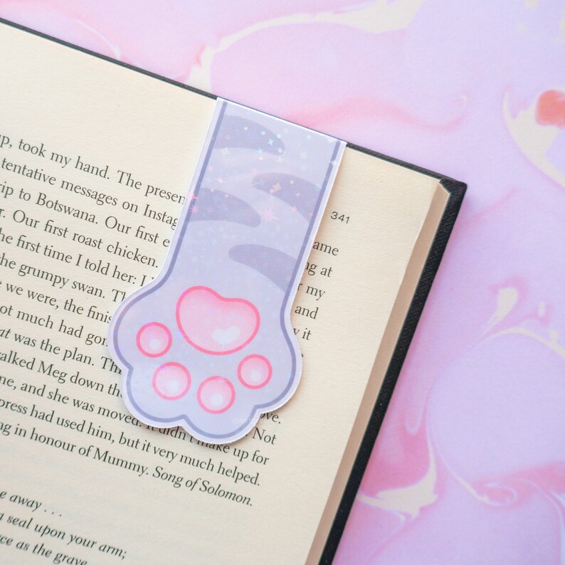 May include: A purple and pink cat paw bookmark with glitter accents. The bookmark is shaped like a cat paw with four toes and a pink pad.