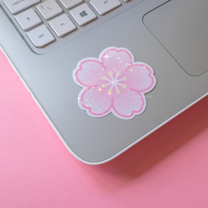 K&ouml;nnte beinhalten: Ein rosa und wei&szlig;er, schimmernder Glitzer-Kirschbl&uuml;ten-Aufkleber auf einem silbernen Laptop.