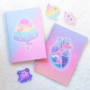 May include: Two pink and purple spiral bound notebooks with colorful illustrations. One notebook features a cotton candy cloud with a pink axolotl and the other features a pink heart with a galaxy inside.