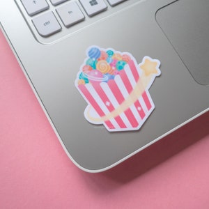 May include: A pink and white striped popcorn bucket filled with colorful planets and a yellow star shooting out of the top. The sticker is on a silver laptop.