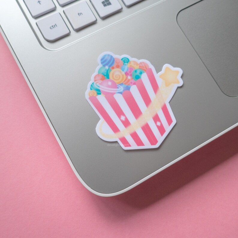May include: A pink and white striped popcorn bucket filled with colorful planets and a yellow star shooting out of the top. The sticker is on a silver laptop.