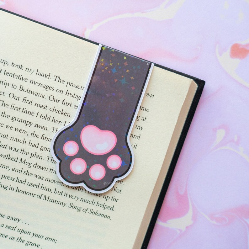 May include: A black and pink paw print bookmark with a holographic background featuring a scattering of stars. The bookmark is made of a durable, flexible material and is perfect for marking your place in a book.