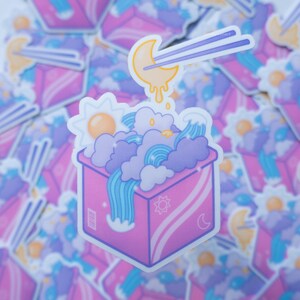 May include: A pink box with a white moon and sun design. The box is filled with blue and purple clouds and a rainbow. A pair of chopsticks with a yellow moon on the end are dipping into the box.