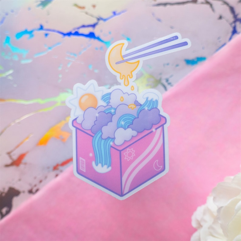 May include: A pink box with a blue, white, and yellow sky inside. The box has a sun and moon design on the side. A yellow crescent moon with a dripping yellow liquid is being lifted by chopsticks.