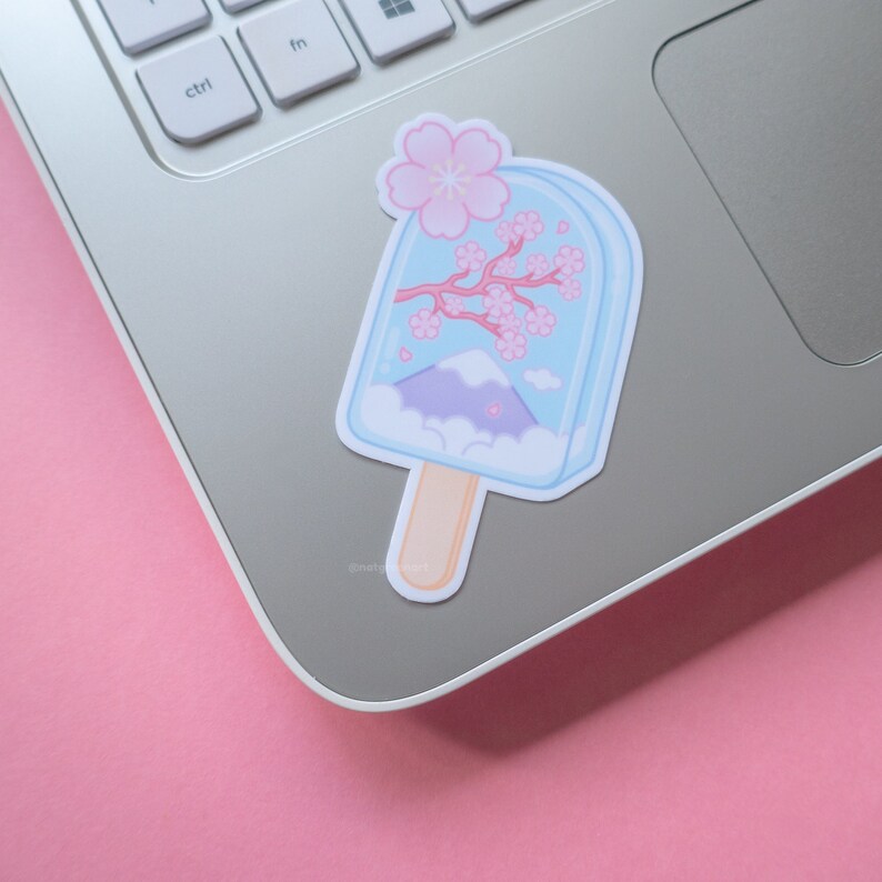 May include: A blue and white popsicle sticker with a pink cherry blossom branch and a mountain scene inside. The sticker is on a silver laptop.