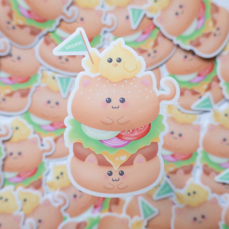 May include: A cute cartoon cat dressed as a vegan burger with a small yellow chick on top. The cat is wearing a green flag that says "VEGAN".
