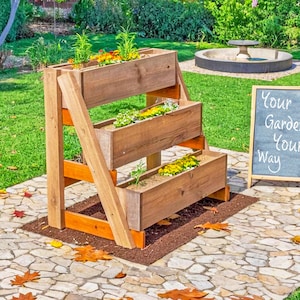 Op de afbeelding: Een houten plantenbak met drie niveaus, gevuld met aarde en planten. De plantenbak is gemaakt van lichtbruin hout en heeft een schuine ondersteuningsstructuur. Een schoolbordbord naast de plantenbak leest "Your Garden Your Way".