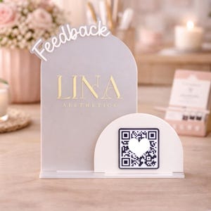 Può includere: Un cartello in acrilico grigio chiaro con la scritta "Feedback" in bianco. Il cartello presenta il testo dorato "LINA Aesthetics" e un codice QR bianco con un cuore. Il cartello è su una superficie di legno.