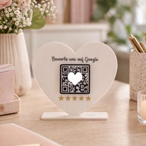 Peut inclure: Un panneau blanc en forme de cœur avec le texte "Bewerte uns auf Google" au-dessus d'un code QR avec un cœur blanc au centre. En dessous du code QR, cinq étoiles dorées. Le panneau est sur une surface en bois.