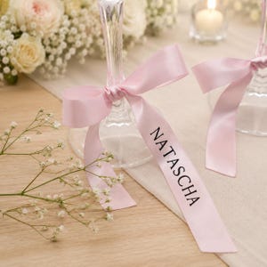 Può includere: Due flûte da champagne adornate con nastri rosa, ognuna con il nome "NATASCHA" in lettere nere. Lo sfondo presenta composizioni floreali e una candela accesa, creando un'atmosfera elegante.