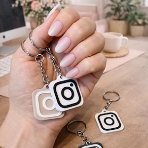 Könnte beinhalten: Schlüsselanhänger mit dem Instagram-Logo in Schwarz, Weiß und Beige. Die Schlüsselanhänger sind quadratisch mit abgerundeten Ecken. Sie sind an silbernen Ketten und Ringen befestigt. Das Bild zeigt mehrere Schlüsselanhänger in einer Hand.