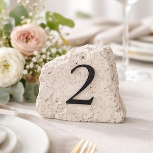 Può includere: Un segnaposto in pietra con il numero 2 in nero. La pietra è di colore beige chiaro e ha una superficie strutturata. Il segnaposto è posto su una tovaglia bianca, con fiori sullo sfondo.