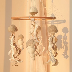 Könnte beinhalten: Ein Holz-Babymobil mit Filz-Meerestieren, die an einem runden Holzrahmen hängen. Das Mobile zeigt weiße und braune Filz-Quallen, Muscheln und Perlen.