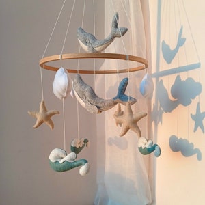 K&ouml;nnte beinhalten: Ein Holz-Babymobil mit maritimem Thema. Es zeigt zwei graue Wale, zwei Seesterne, zwei Wolken und zwei Wellen, alles aus Filz. Das Mobile h&auml;ngt an einer wei&szlig;en Decke.